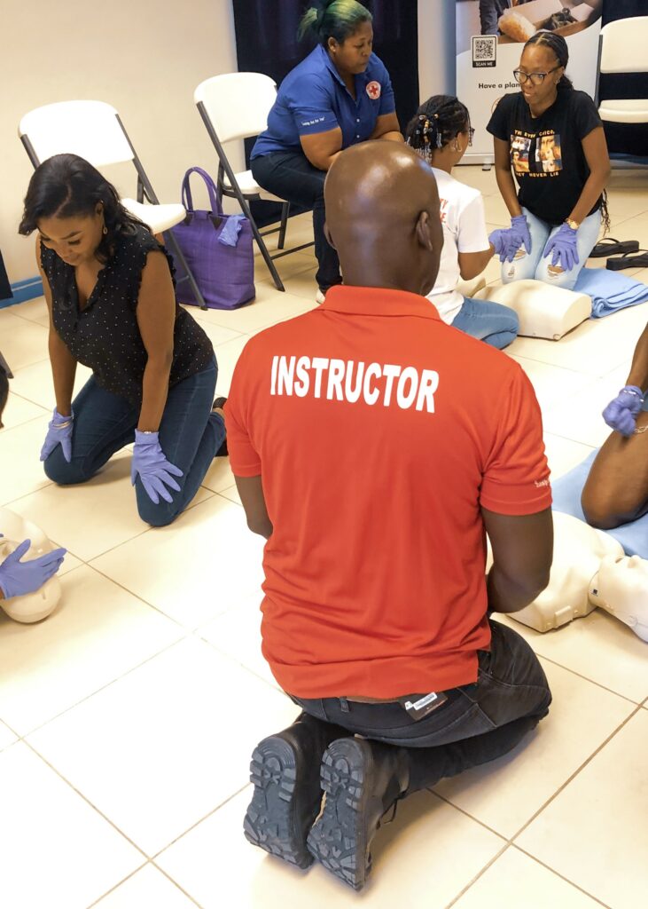 Image of a man’s back as he kneels while instructing a group of women in applying CPR.